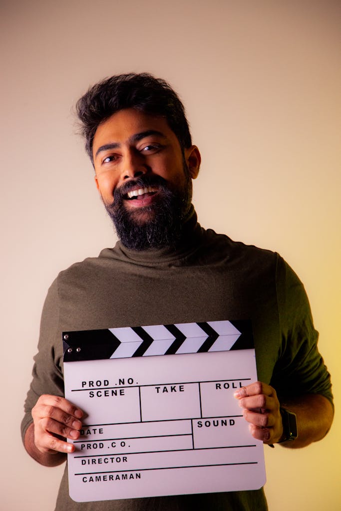 A smiling man with a beard holding a film clapboard in a studio setting, ready for movie production.