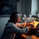 A thoughtful writer smokes a cigarette while working at a vintage desk, illuminated by moody lighting.